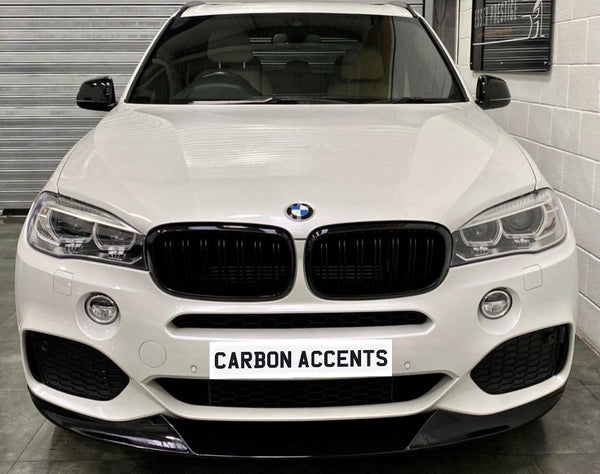 A white BMW SUV is parked indoors, facing forward with a sign on the front reading "CARBON ACCENTS." Surrounding are a tiled floor and walls with metal shuttering.