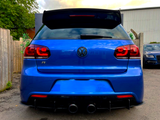 A blue Volkswagen car, parked with visible dual exhausts and a spoiler, is situated between a wooden fence and a metal building, with trees and another car nearby.