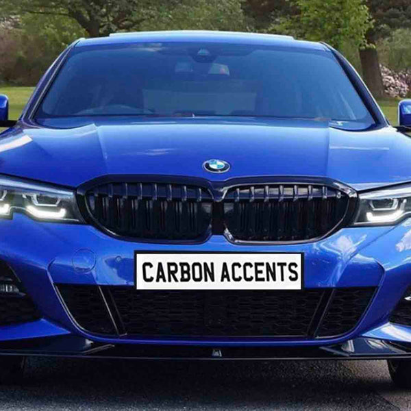 A blue car with illuminated headlights is parked on a road within a park-like setting. The license plate reads, "CARBON ACCENTS."