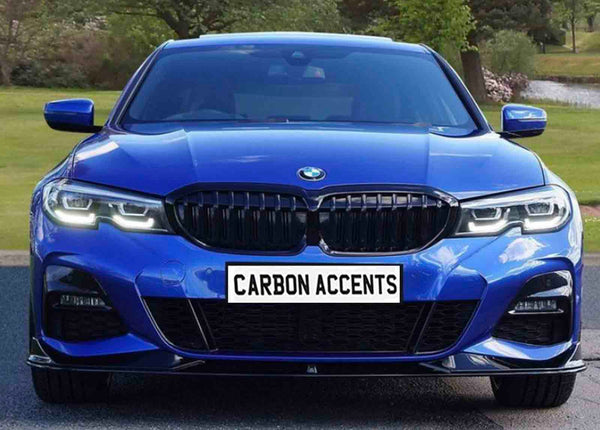 A blue car with illuminated headlights is parked on a road within a park-like setting. The license plate reads, "CARBON ACCENTS."