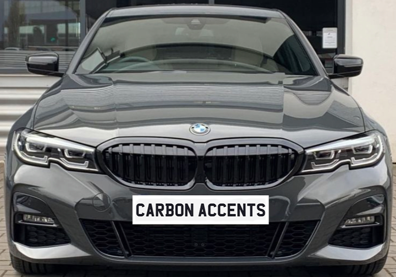 A gray BMW car is parked, facing forward with sleek design elements. Text on the front reads "CARBON ACCENTS." It's positioned outdoors on a brick surface.