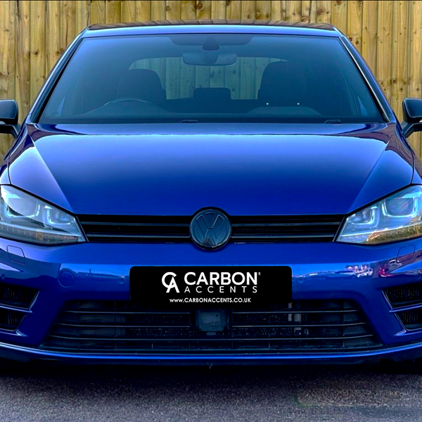 A blue car is parked facing forward, displaying the logo on its grille. "CARBON ACCENTS" is on the license plate. The vehicle is set against a wooden fence background.