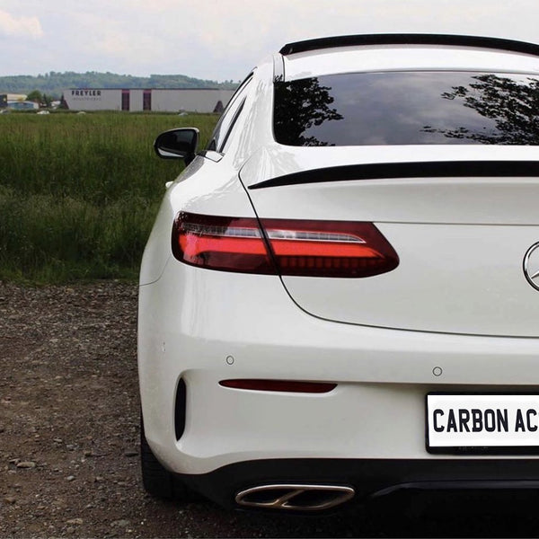 A white Mercedes-Benz car is parked on gravel, facing away. The license plate reads "CARBON ACCENT." In the background, a grassy field and industrial buildings are visible.