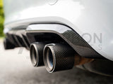 Exhaust pipes with carbon fiber surround, protruding from a vehicle's rear, parked on asphalt with blurred foliage in the background.