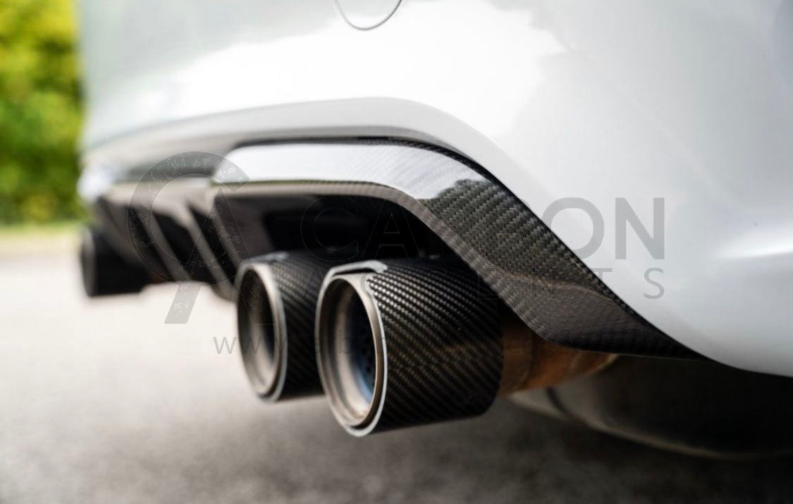 Exhaust pipes with carbon fiber surround, protruding from a vehicle's rear, parked on asphalt with blurred foliage in the background.