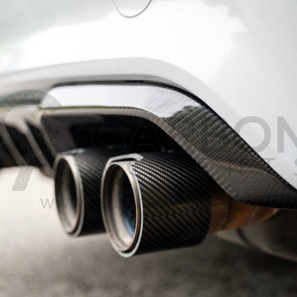 Exhaust pipes with carbon fiber surround, protruding from a vehicle's rear, parked on asphalt with blurred foliage in the background.