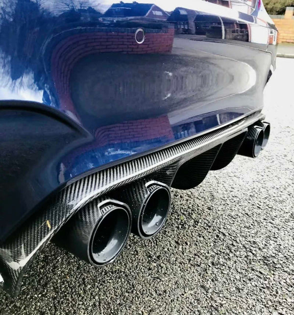 Car exhaust pipes, featuring four round outlets framed by carbon fiber, are situated on a dark car's rear metallic bumper. Parked on a textured asphalt surface, with brick walls reflected.