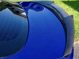 A blue car with a carbon fiber spoiler reflects sunlight, parked on grass. The rear window shows building reflections, emphasizing the car's glossy finish and sporty design.