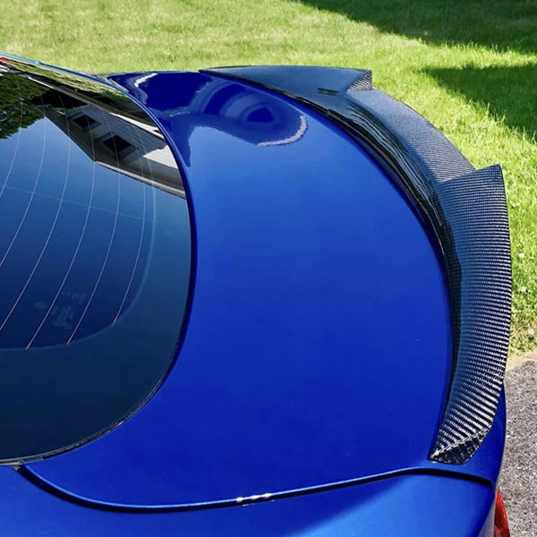 A blue car with a carbon fiber spoiler reflects sunlight, parked on grass. The rear window shows building reflections, emphasizing the car's glossy finish and sporty design.