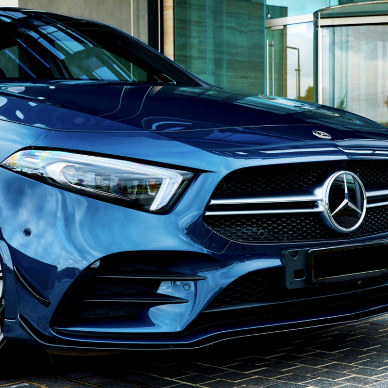 A blue Mercedes-Benz car is parked on a cobblestone surface, next to a modern glass building entrance. The focus is on the front grille and headlight details.