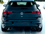 A black Volkswagen car is parked on a snow-covered road in a forested area, showing dual exhaust pipes and illuminated tail lights.