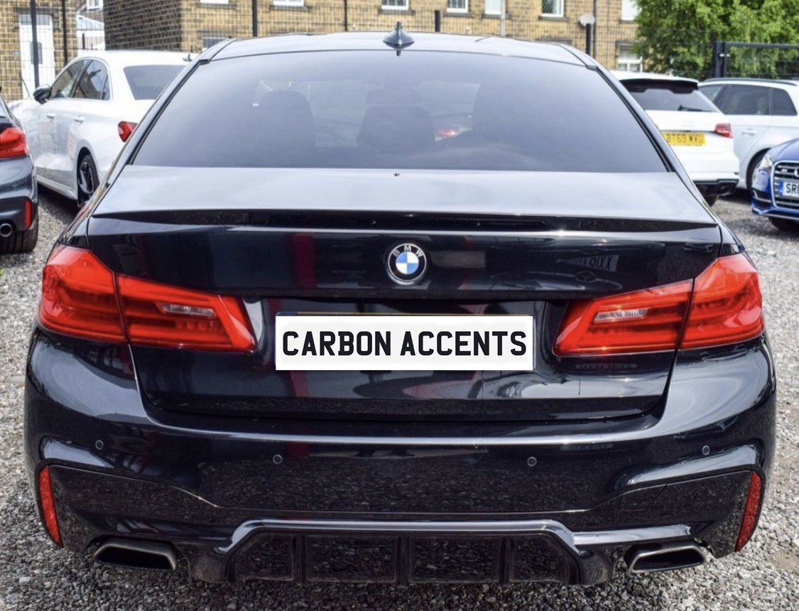 A black BMW with red tail lights is parked on gravel in a lot, surrounded by other vehicles. The license plate reads "CARBON ACCENTS."