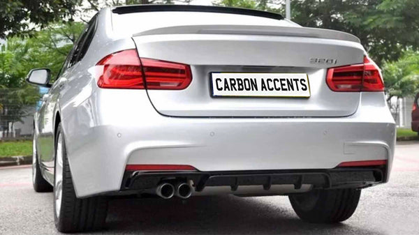 A silver car is parked on a paved surface with greenery in the background. The rear features dual exhausts and a license plate reading "CARBON ACCENTS."