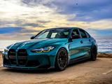 A sleek, blue sports sedan is parked beside the ocean on a sandy surface, under a dramatic, cloudy sky at sunset.
