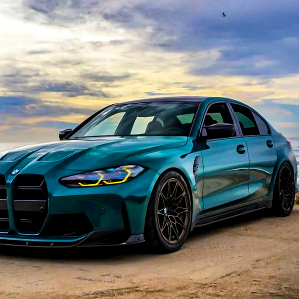 A sleek, blue sports sedan is parked beside the ocean on a sandy surface, under a dramatic, cloudy sky at sunset.