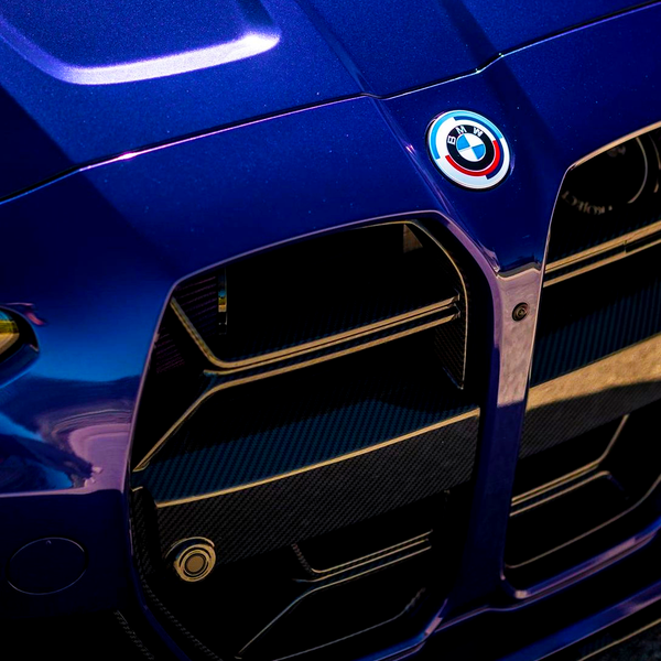 A blue car front displays a prominent grille and a round BMW emblem above it, situated outdoors in bright sunlight.