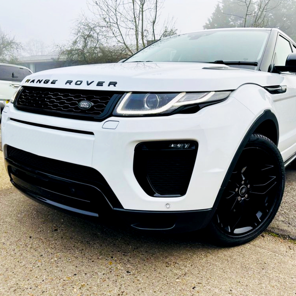 A white Range Rover parked on a concrete surface with visible branding. It is surrounded by other vehicles on a foggy day, with trees and fencing in the background.