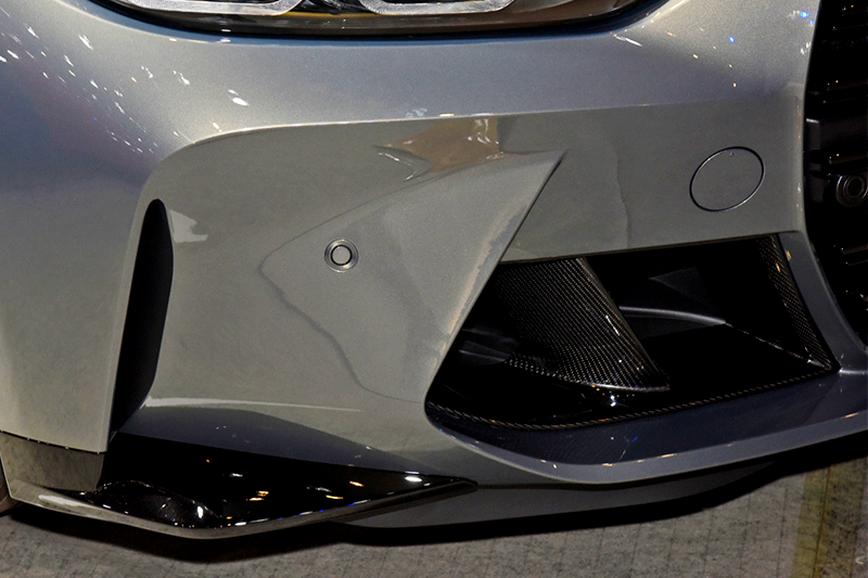 A sleek car bumper, featuring a metallic silver finish, rests motionless. It includes a circular sensor and black air vents, set in a showroom with overhead lighting reflections.