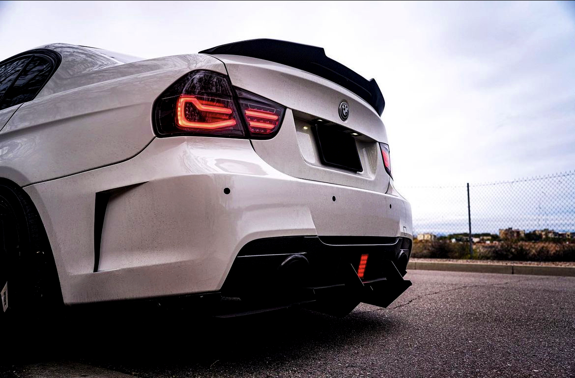A white sports car with illuminated red taillights is parked on a paved road, near a fence, under a cloudy sky.