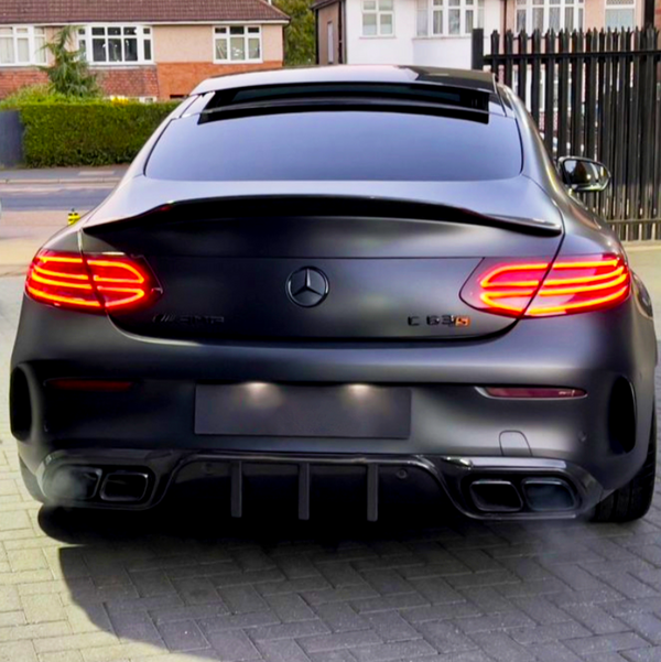 A black Mercedes-Benz C 63 S AMG is parked, with illuminated tail lights, on a paved driveway. Houses and a black metal fence are visible in the background.