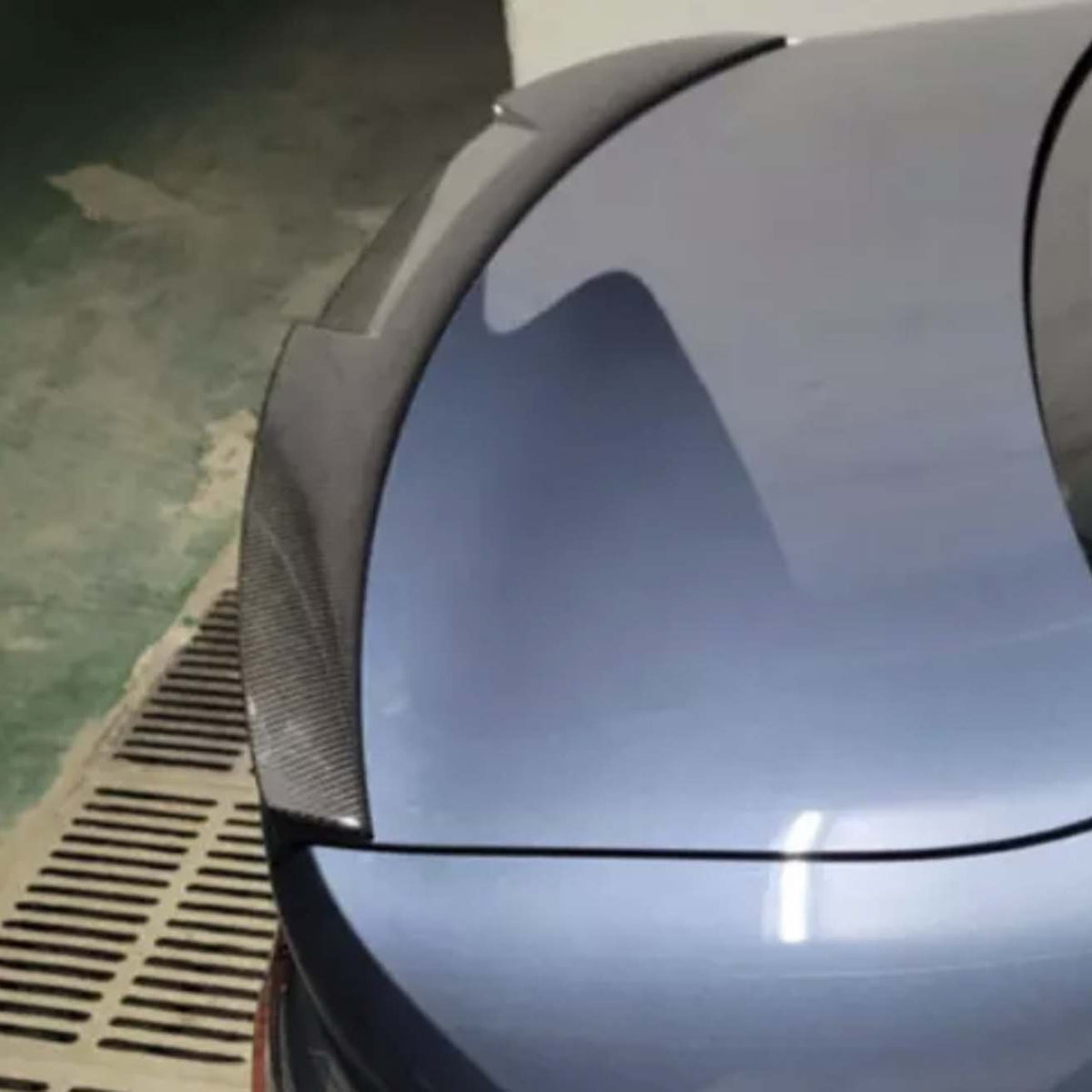A car trunk with a carbon fiber spoiler is parked in an indoor garage with a green and beige floor, next to a drainage grate.