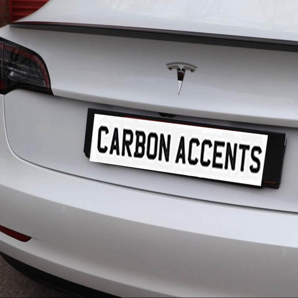 A white car's rear view shows a "CARBON ACCENTS" license plate, with a Tesla logo above it, parked on a concrete surface.