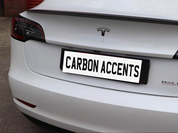 A white car's rear view shows a "CARBON ACCENTS" license plate, with a Tesla logo above it, parked on a concrete surface.