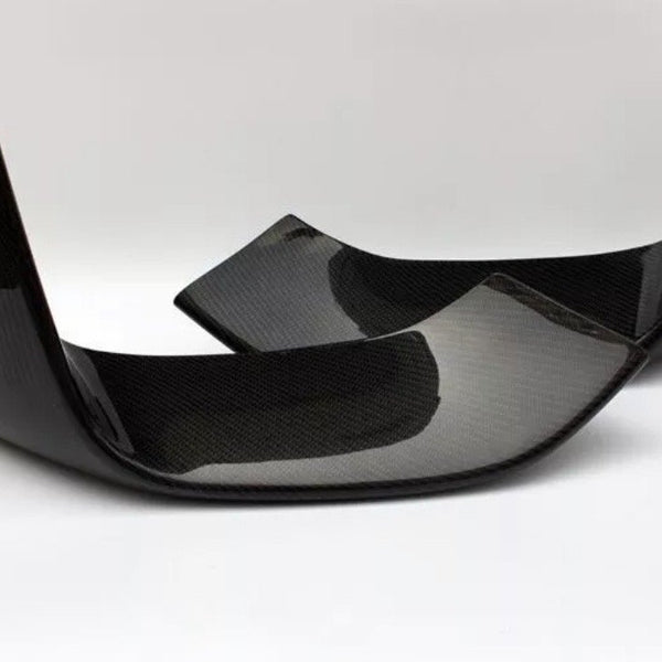 Two carbon fiber car parts, possibly rear exhaust diffusers, are positioned diagonally on a solid white surface, highlighting their glossy black finish and textured pattern.