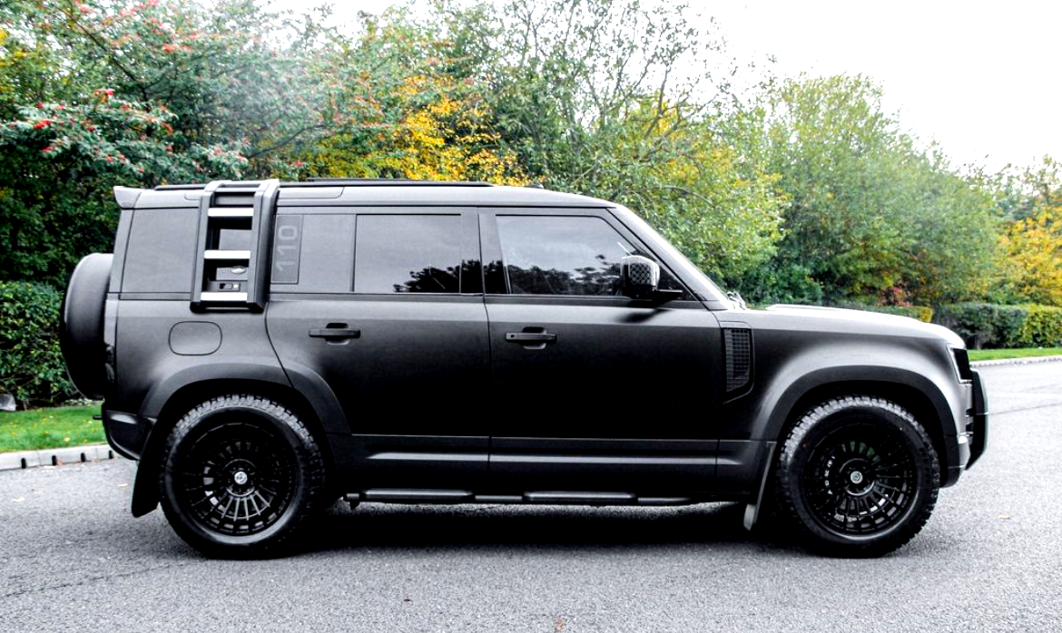 A black SUV is parked on a road, featuring black wheels and tinted windows, with a lush, green, and slightly autumnal forest backdrop.