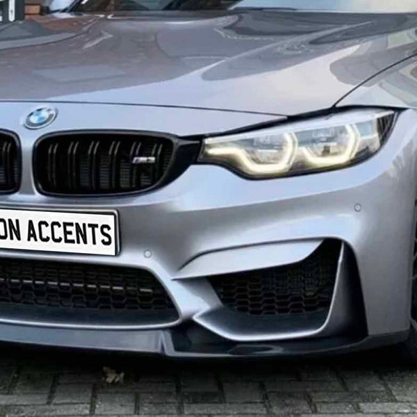 A sleek, grey sports car is parked on a brick pavement, displaying a license plate that reads "CARBON ACCENTS." The headlights are illuminated, and the front grille is prominent.