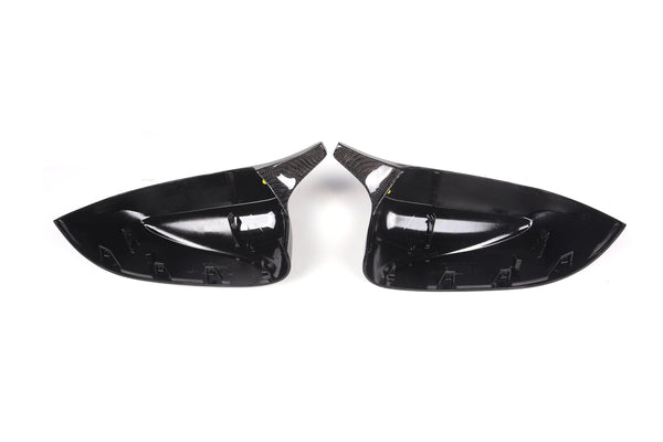 Two black, glossy car side mirror covers face each other, highlighting their contoured interior and carbon fiber textures, set against a plain white background.