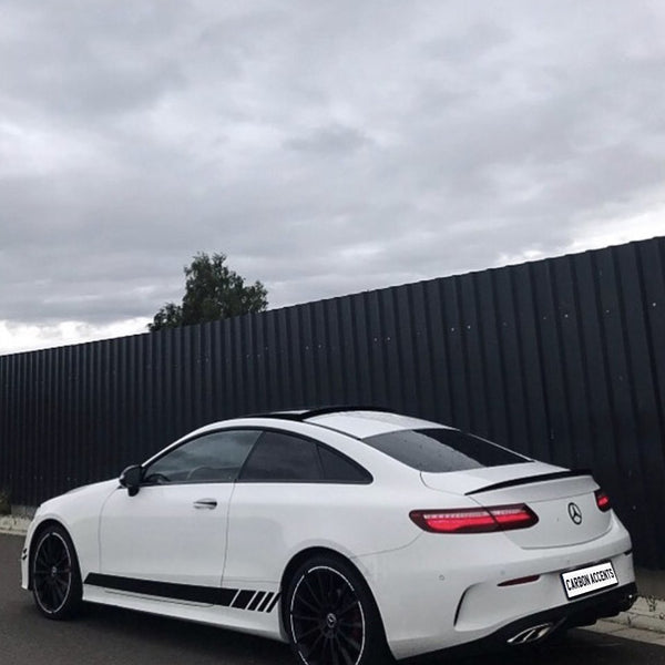 A white sports coupe is parked on a street beside a tall, dark fence under a cloudy sky. The license plate reads "CARBON ACCENTS."