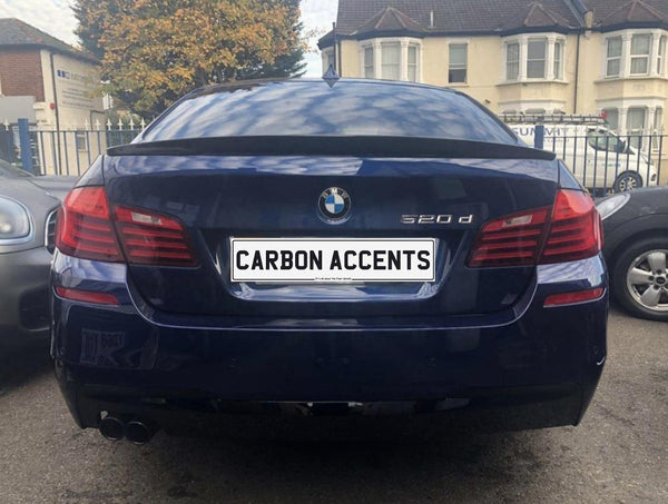 A blue BMW 520d is parked with its rear facing the camera. The license plate reads "CARBON ACCENTS." Nearby are other cars, buildings, and a tree.