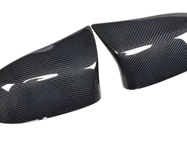 Two carbon fiber mirror covers rest side by side, reflecting light on their glossy surface, set against a plain white background.
