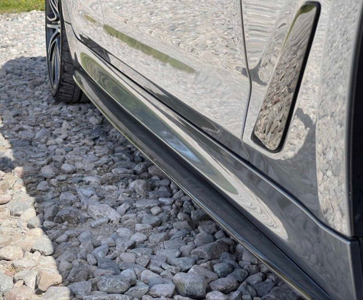 A car's sleek, reflective side is parked on a gravel surface, showcasing its glossy finish and detailed wheel rims in a sunlit environment.