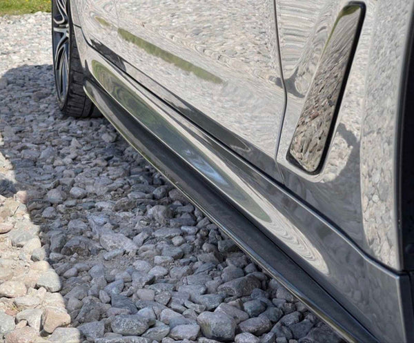 A car's sleek, reflective side is parked on a gravel surface, showcasing its glossy finish and detailed wheel rims in a sunlit environment.