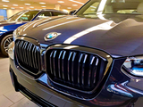 The front of a dark-colored car with a prominent grille and BMW logo is parked indoors alongside other vehicles under bright ceiling lights.