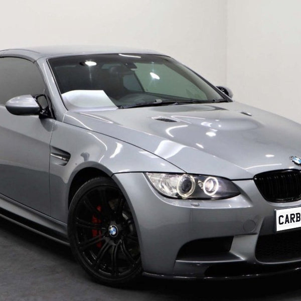 A sleek, silver sports car with tinted windows and black rims sits in a white showroom. The license plate reads "CARBON ACCENTS."