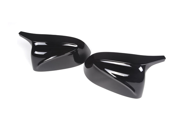Two glossy black aerodynamic mirror covers lie on a white surface, their sleek, curved design reflecting light, indicating they are automotive accessories.