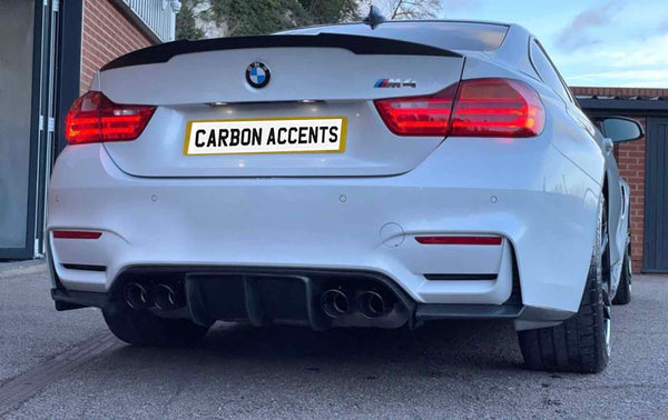 A white BMW M4 displays illuminated red taillights. The license plate reads "CARBON ACCENTS." The car is parked near a building with red brick and a gray door.