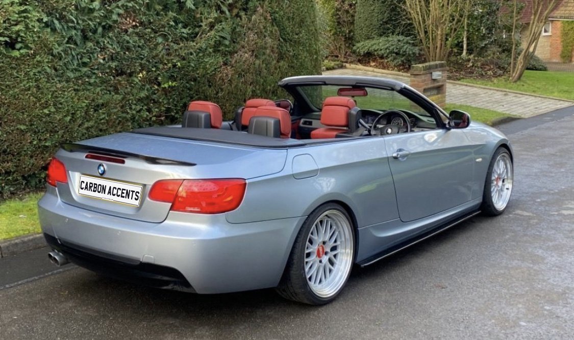 Convertible car with red interior parked on a driveway. License plate reads "CARBON ACCENTS." Surrounding environment includes greenery and a brick house in the background.