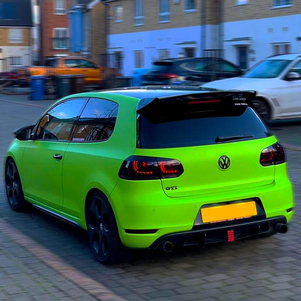 A bright green Volkswagen hatchback is parked in a residential street, displaying "GTI" on the rear. Other parked cars are visible, with brick houses lining the background.