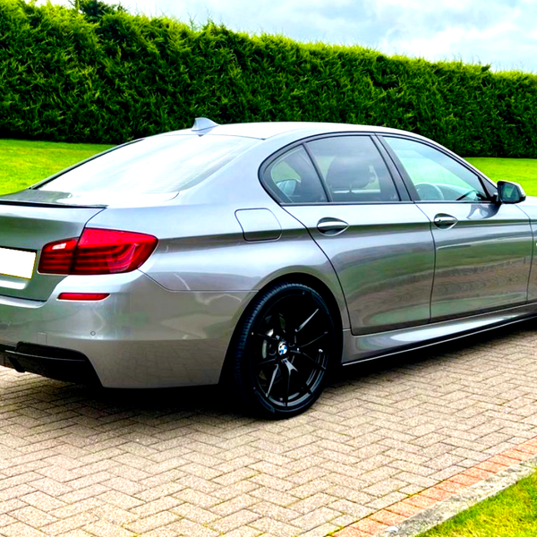 A silver sedan is parked on a brick driveway. It has black wheels and red rear lights. The background features a green lawn and tall hedges under a cloudy sky.
