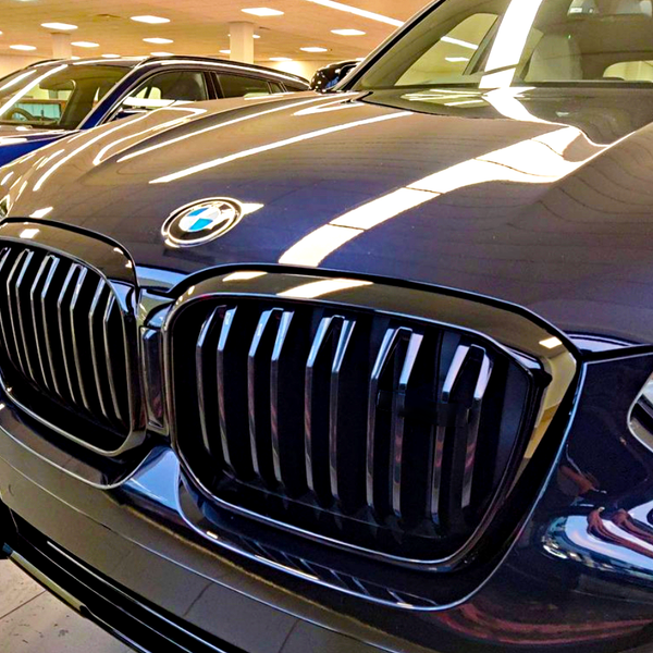 A shiny BMW car sports a prominent grille in a well-lit showroom, surrounded by other vehicles and illuminated by overhead lights.