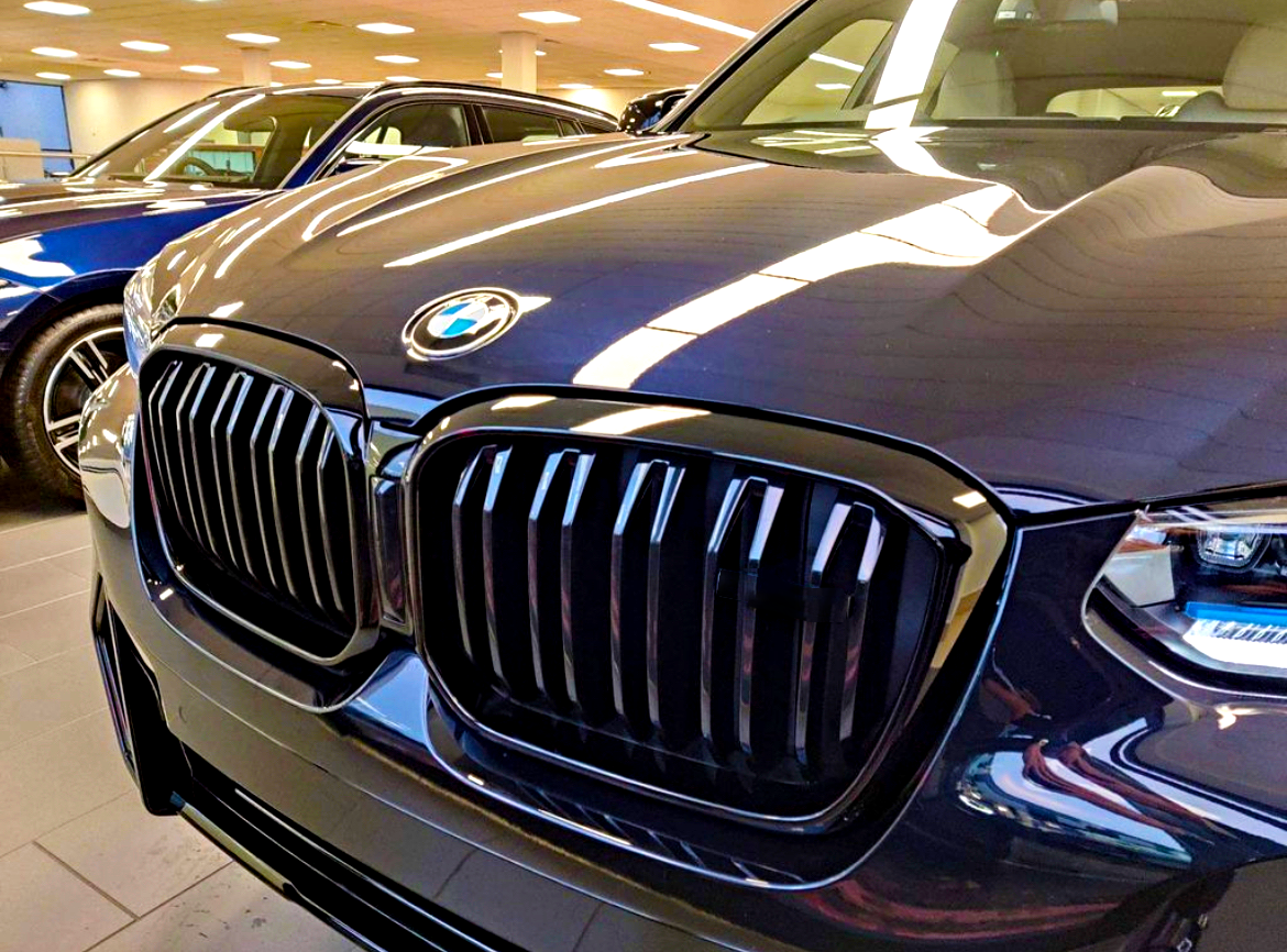 A shiny BMW car sports a prominent grille in a well-lit showroom, surrounded by other vehicles and illuminated by overhead lights.