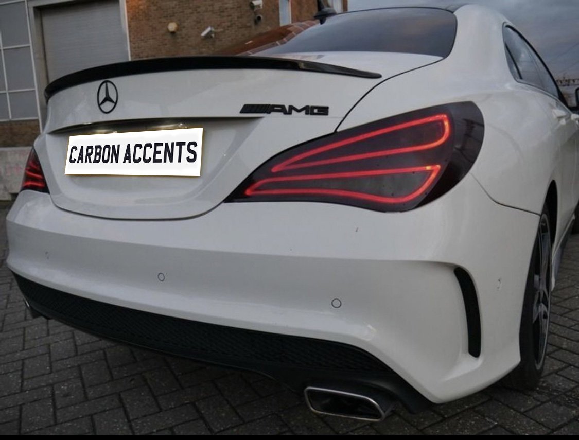 A white car with a Mercedes emblem and AMG badge is parked on a paved surface. The license plate reads, "CARBON ACCENTS." The brick building is visible in the background.