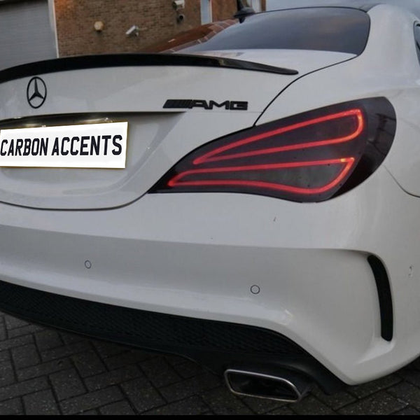 A white car with a Mercedes emblem and AMG badge is parked on a paved surface. The license plate reads, "CARBON ACCENTS." The brick building is visible in the background.