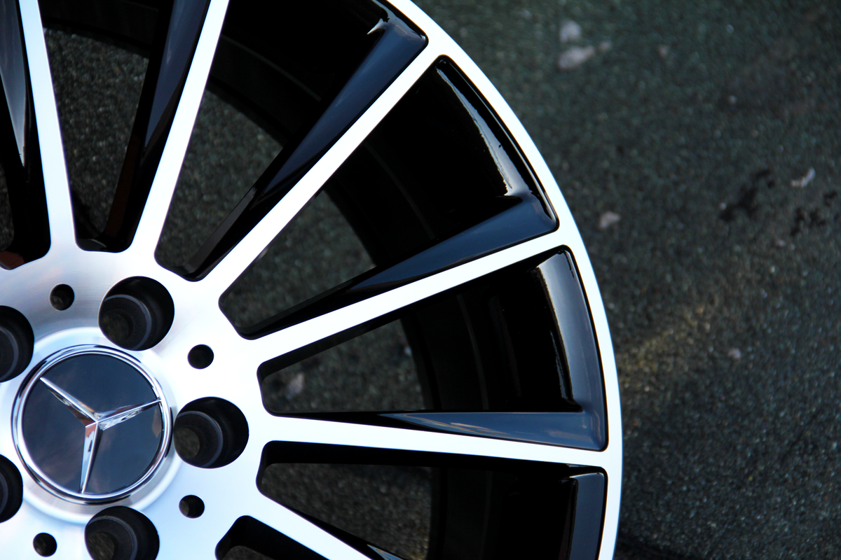 Wheel rim with a metallic Mercedes-Benz logo and black spokes, resting still, set against a textured asphalt surface.