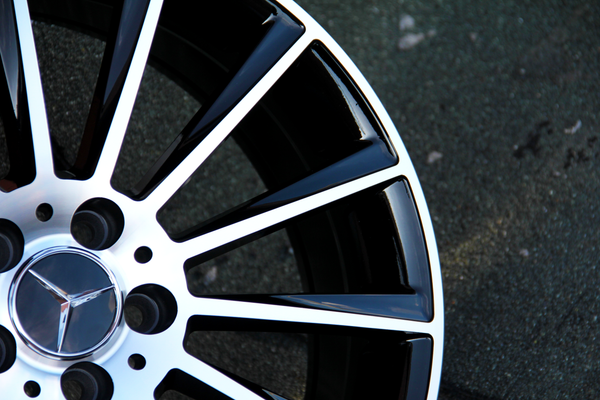 Wheel rim with a metallic Mercedes-Benz logo and black spokes, resting still, set against a textured asphalt surface.