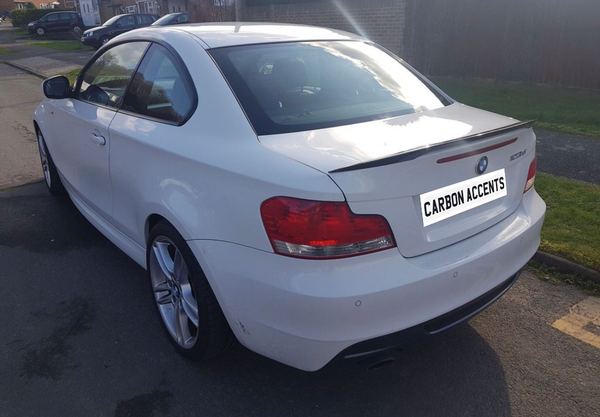 A white BMW 123d, parked on a street, displays a carbon spoiler. Text on the rear reads "CARBON ACCENTS." Other cars and houses are visible in the background.